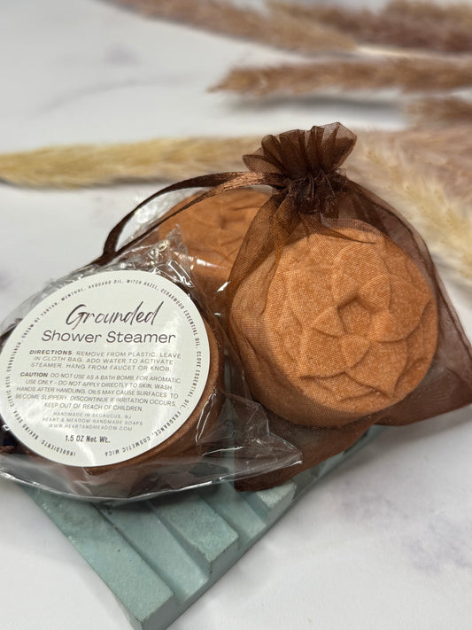 A brown shower steamer in a sheer brown drawstring bag with a clear plastic wrap and a round white label reading 'Grounded Shower Steamer,' placed on a light surface with dried grasses in the background.