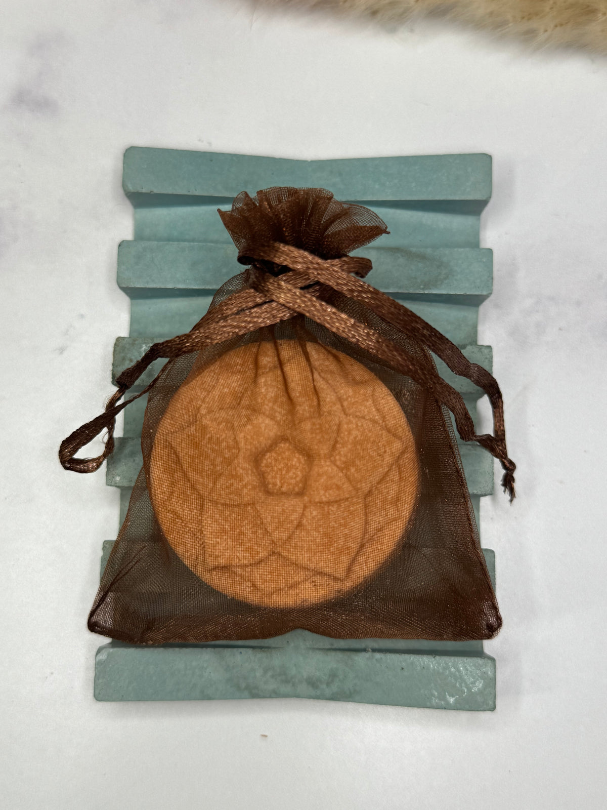 A round tan shower steamer with an embossed geometric lotus design, sealed in a brown sheer organza drawstring pouch and displayed on a light blue wooden stand against a white marble background.