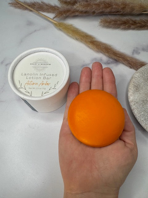 An orange-colored lanolin-infused lotion bar held in a person's hand, with its white container labeled 'Heart & Willow Lotion Bar' on a light marble surface with dried grasses in the background.